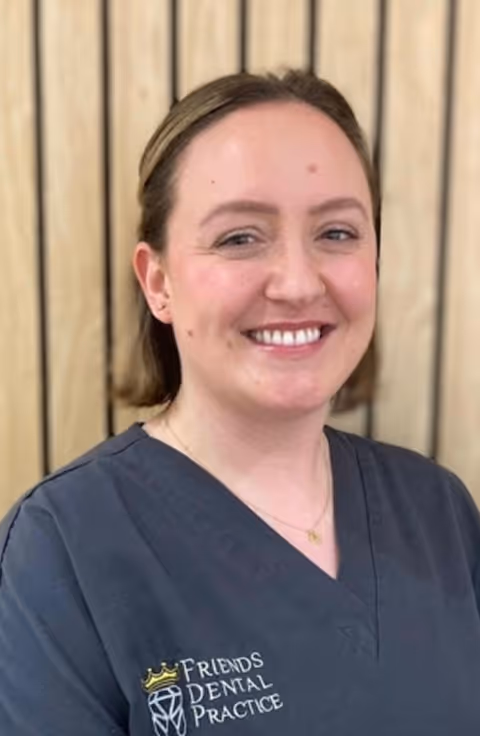 Smiling woman wearing a dark gray dental uniform with Friends Dental Practice logo, standing in front of a wooden panel background.
