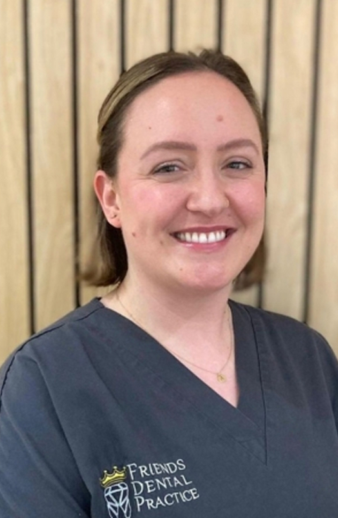 Smiling woman wearing a dark gray dental uniform with Friends Dental Practice logo, standing in front of a wooden panel background.