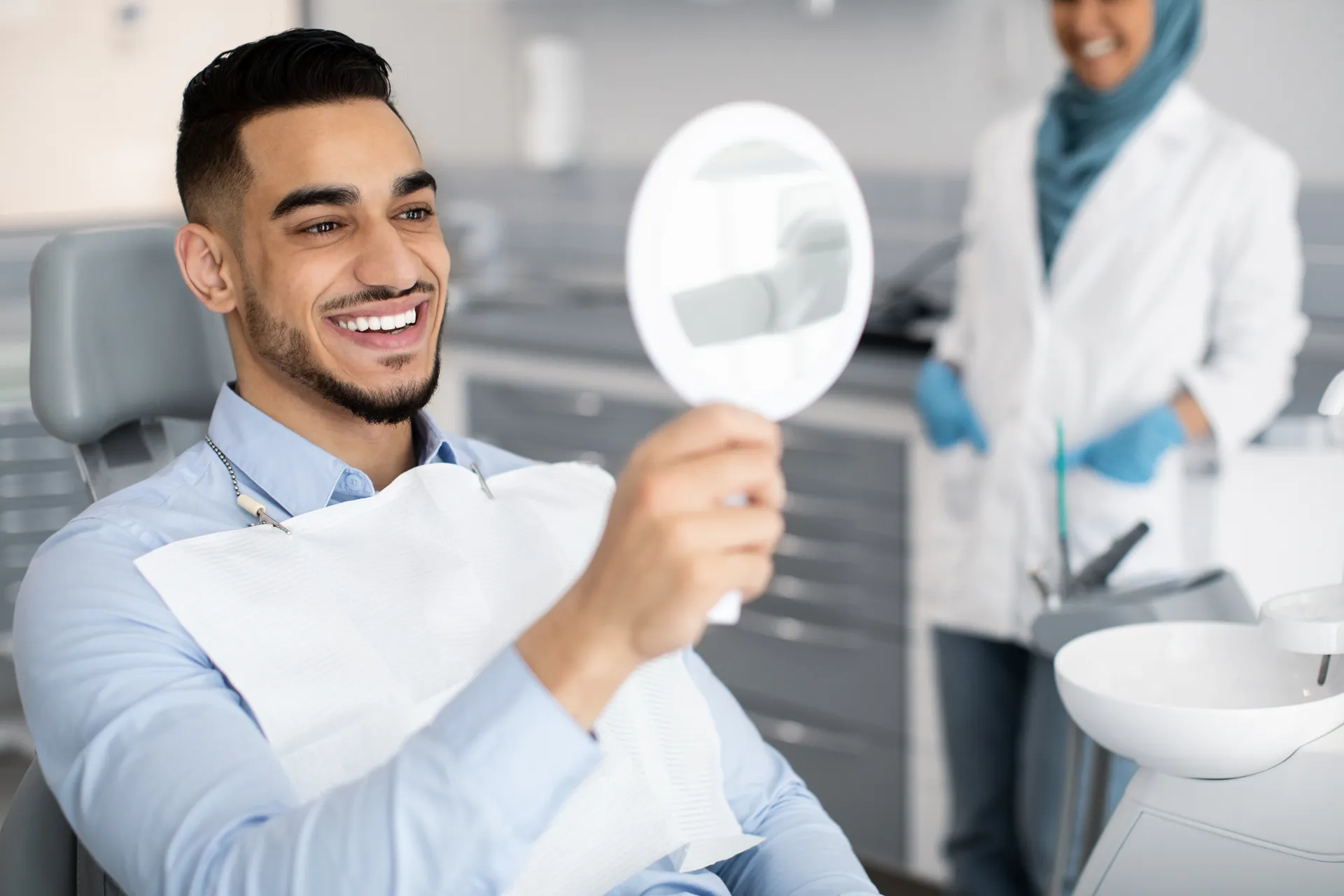 Smiling man in dental chair holding a mirror, with a dentist in a white coat and blue gloves in the background.