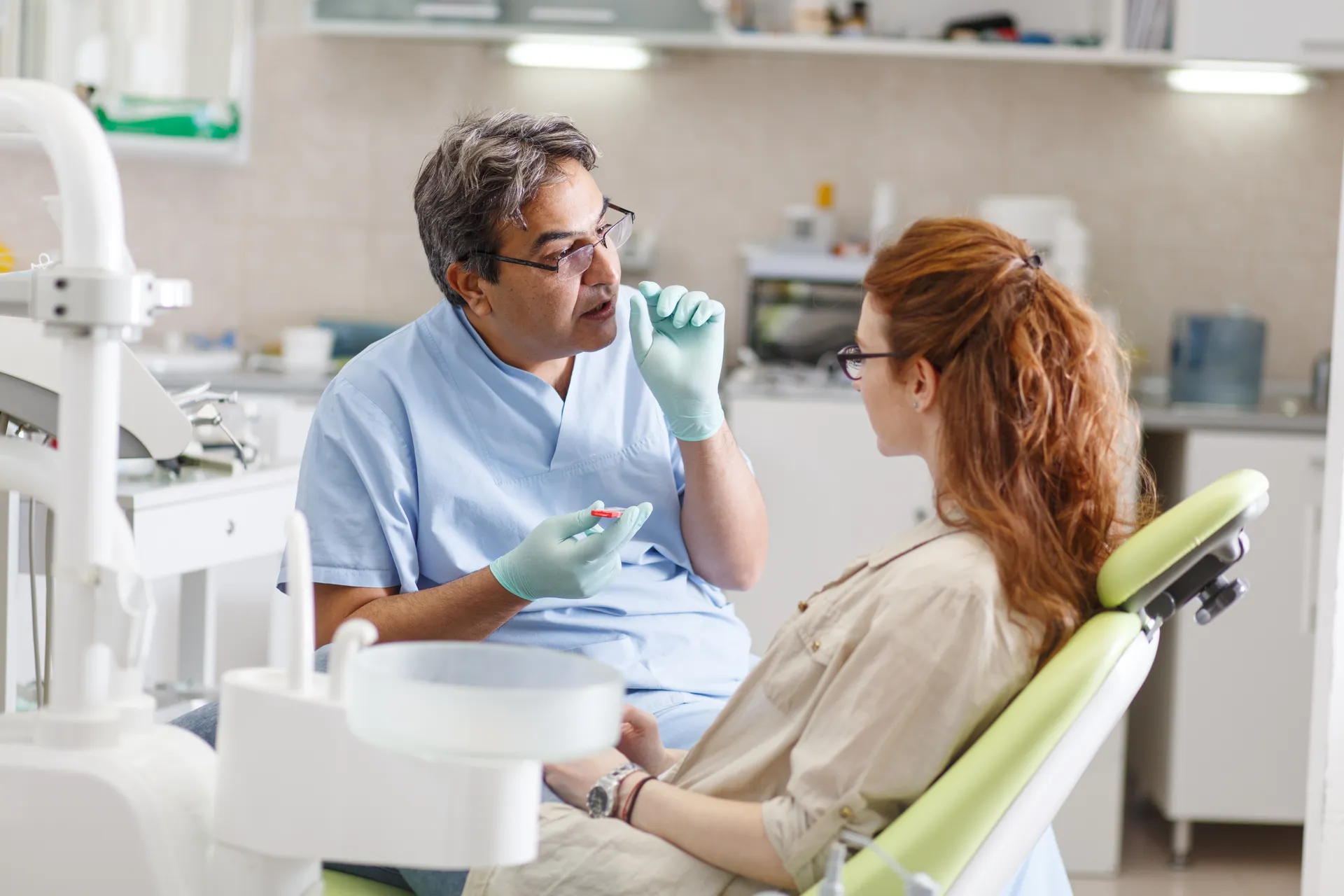 Dentist in blue scrubs explaining dental care to a female patient seated in a dental chair.