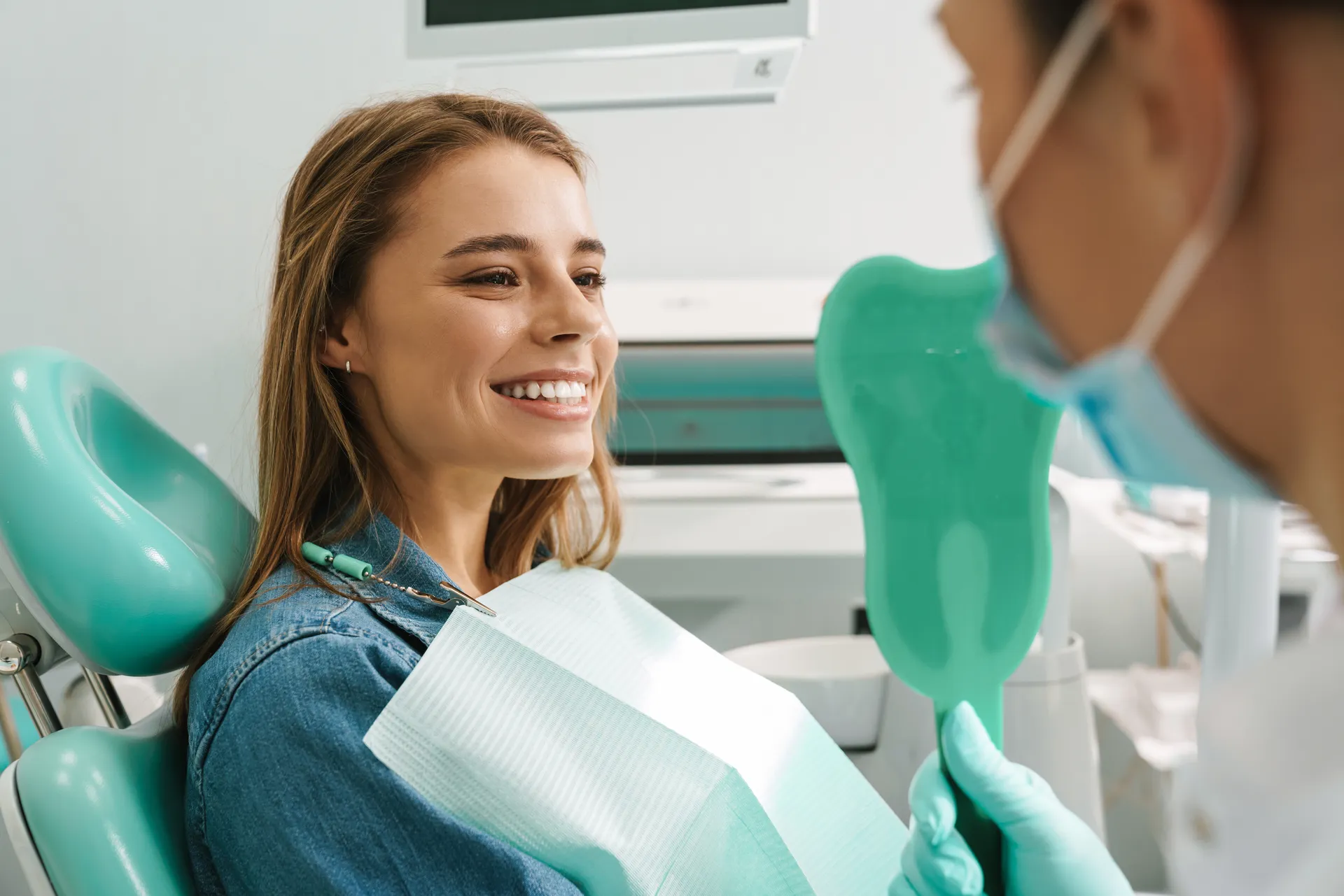 Smiling woman sitting in a dental chair looking at her teeth in a tooth-shaped mirror held by a dentist wearing a mask.