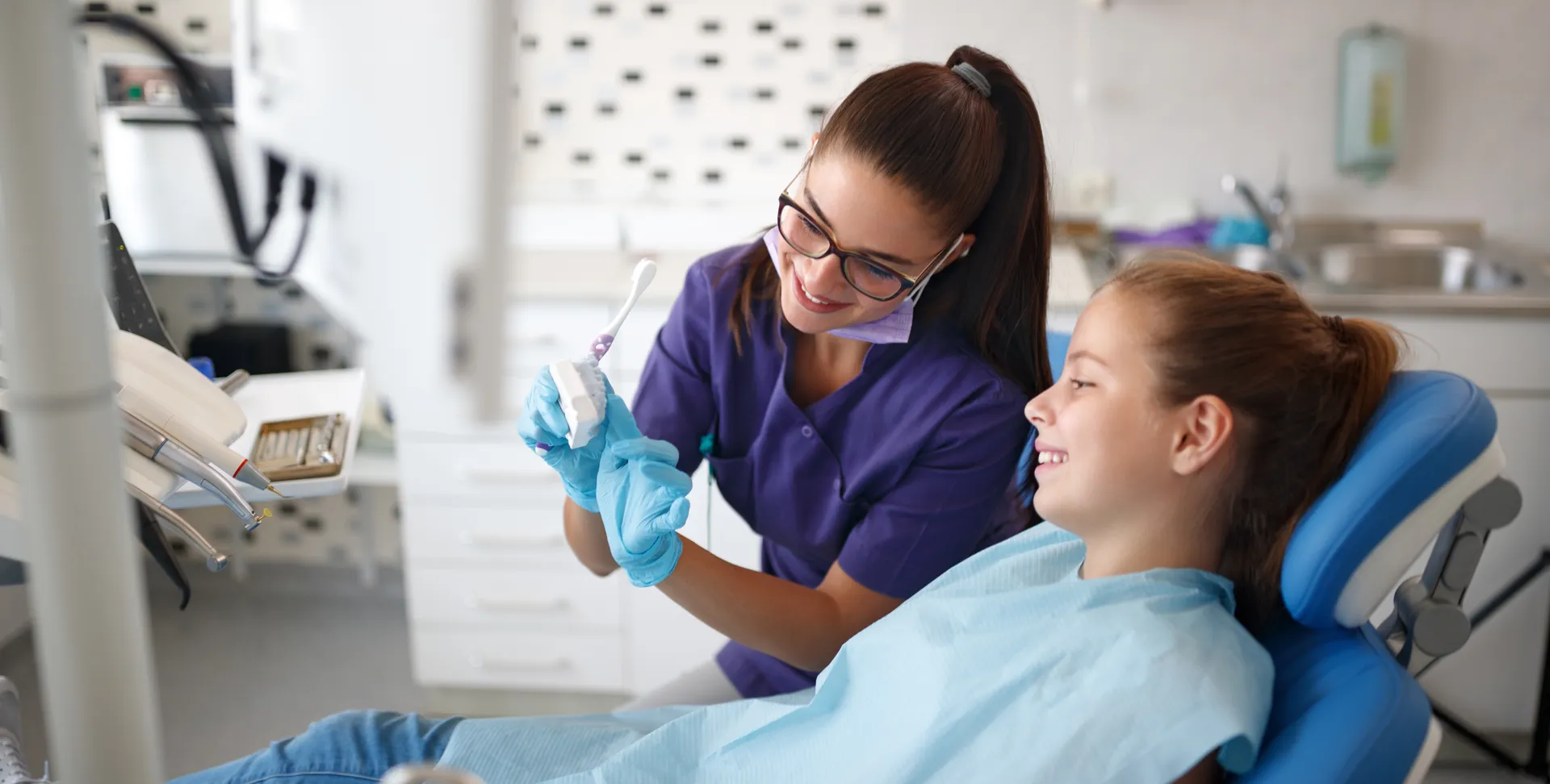 Dental hygienist in purple scrubs demonstrating toothbrushing technique using a dental model to a smiling young girl in a dental chair.