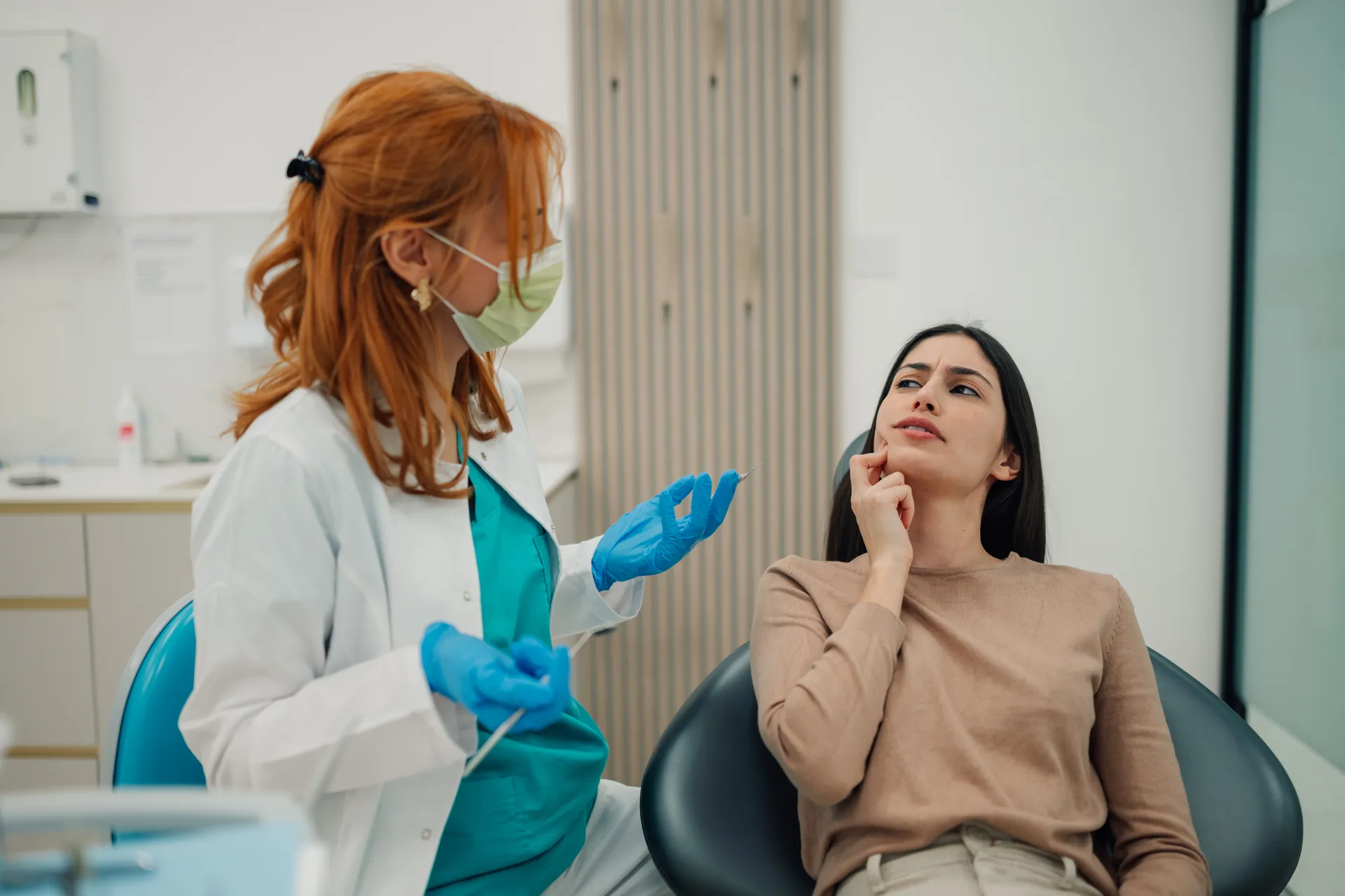 Female dentist wearing a mask and gloves talking to a woman patient who is pointing to her jaw in discomfort.