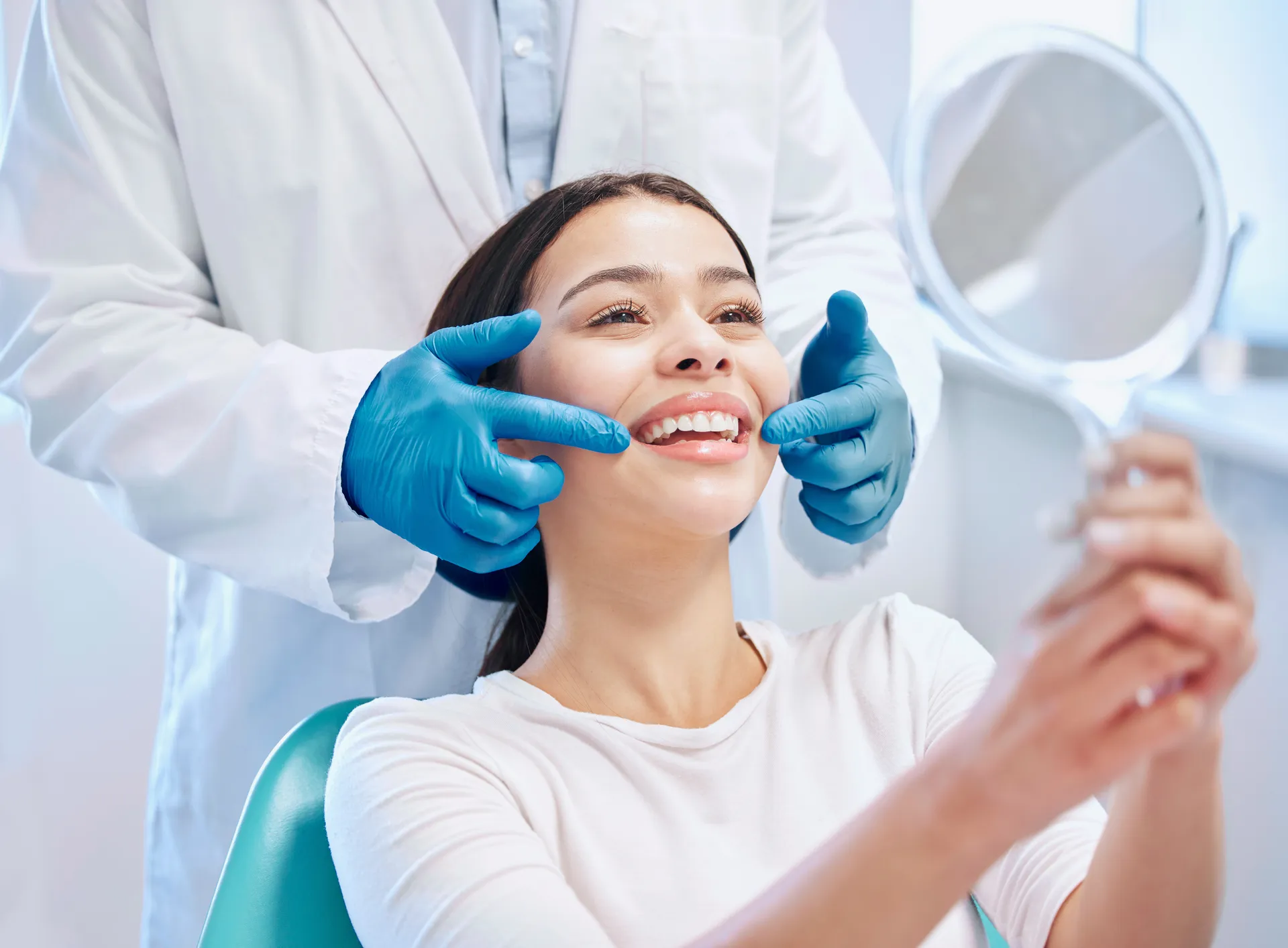 Smiling woman in a dental chair holding a mirror while a dentist wearing blue gloves examines her teeth.