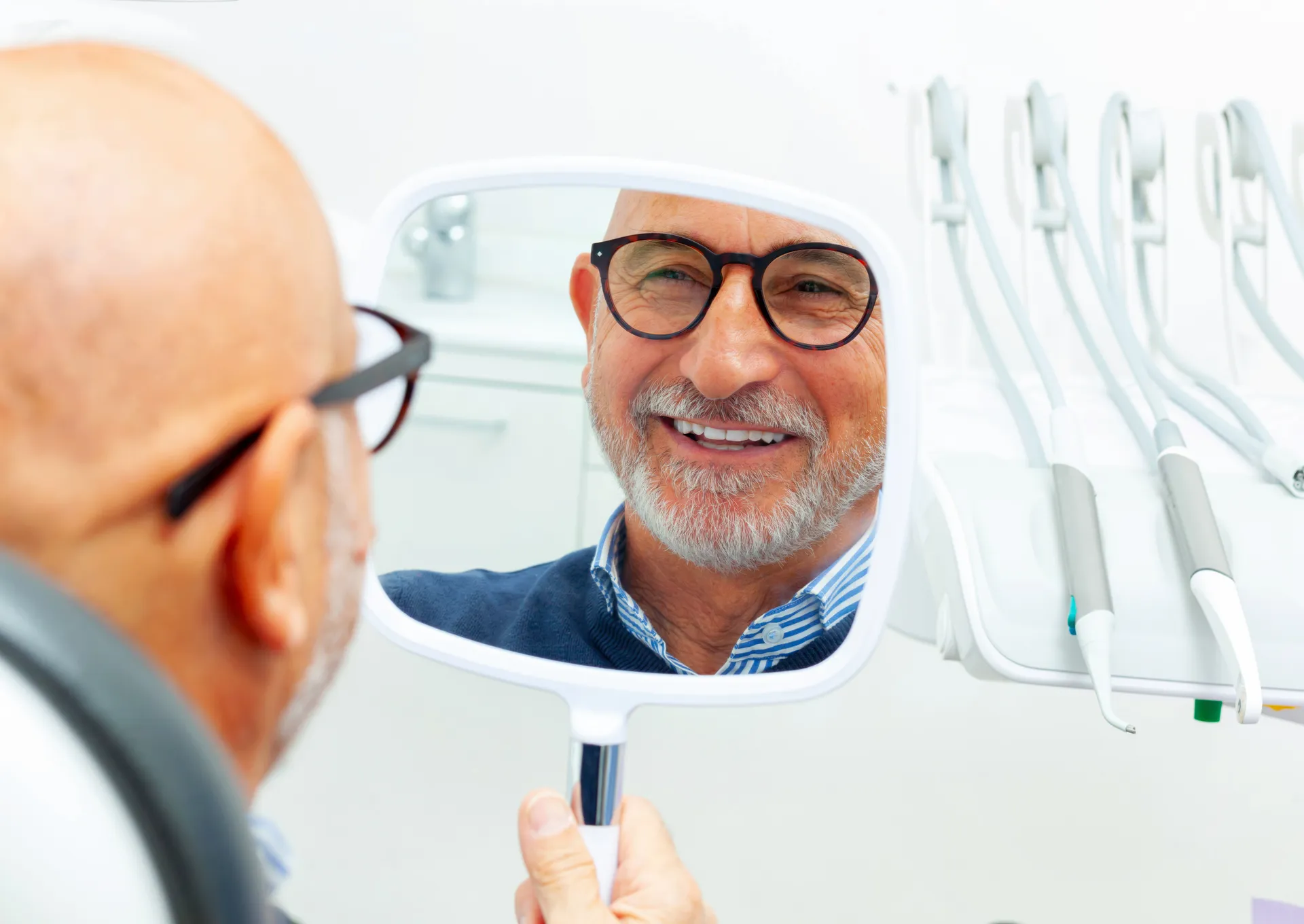 Smiling elderly man with glasses holding a handheld mirror in a dental clinic, checking his teeth.