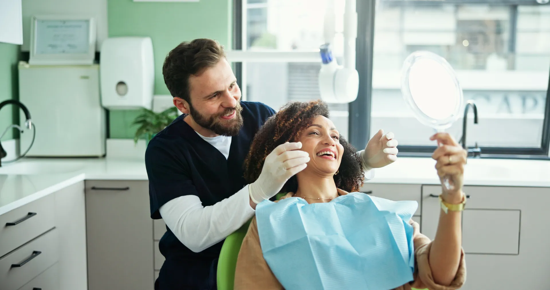 Dentist smiling and interacting with a female patient who is looking at her teeth in a handheld mirror.