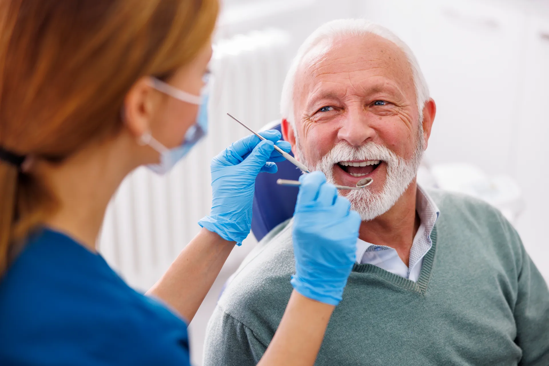 Dentist wearing blue gloves and mask examining an elderly man's teeth in a dental clinic.