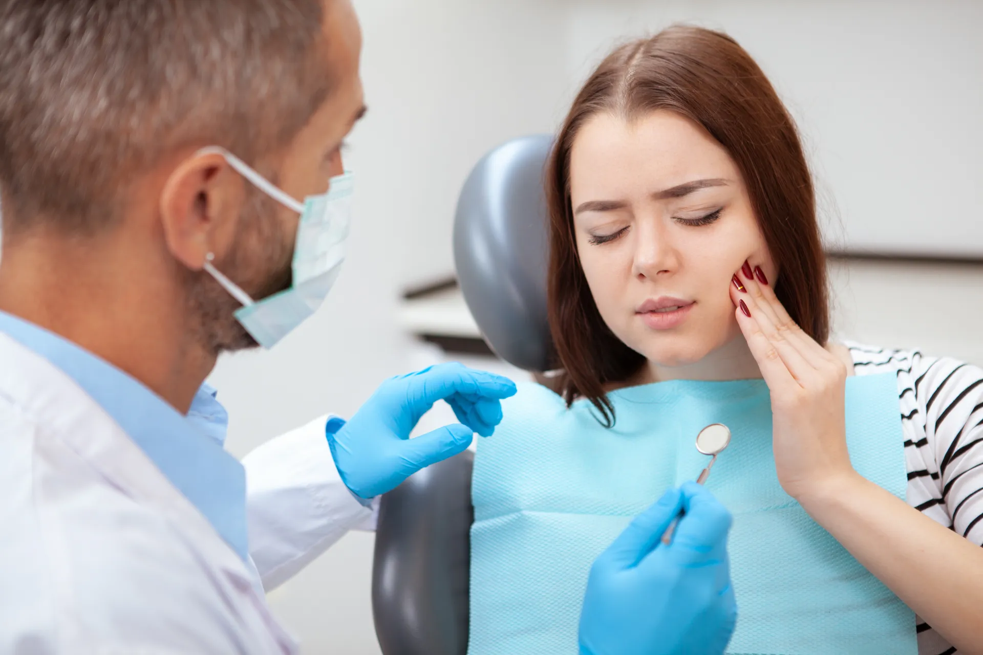 Young woman in dental chair holding her cheek in pain as dentist wearing mask and gloves holds a dental mirror.