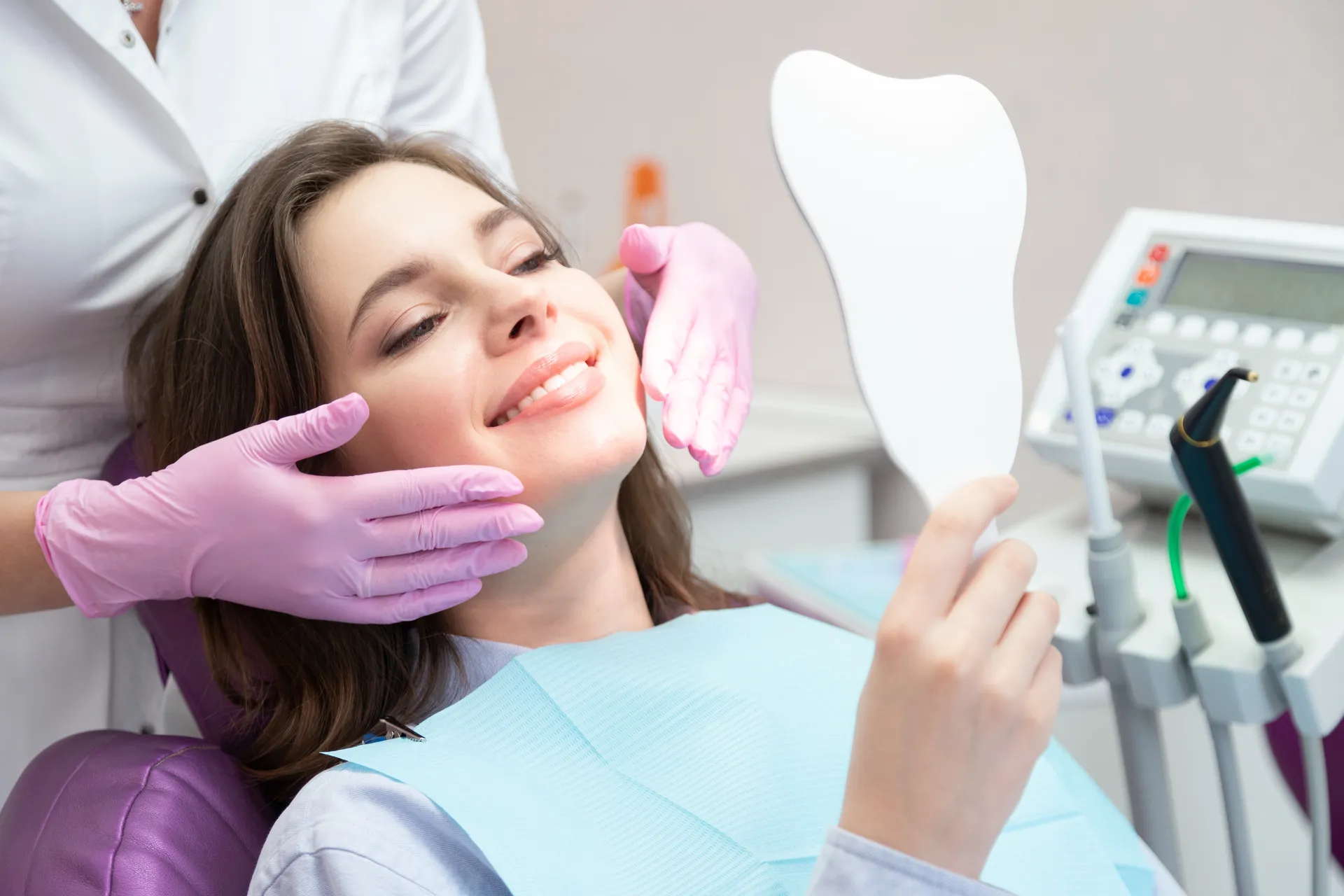 Smiling woman sitting in dental chair holding a tooth-shaped mirror while dentist wearing pink gloves examines her face.