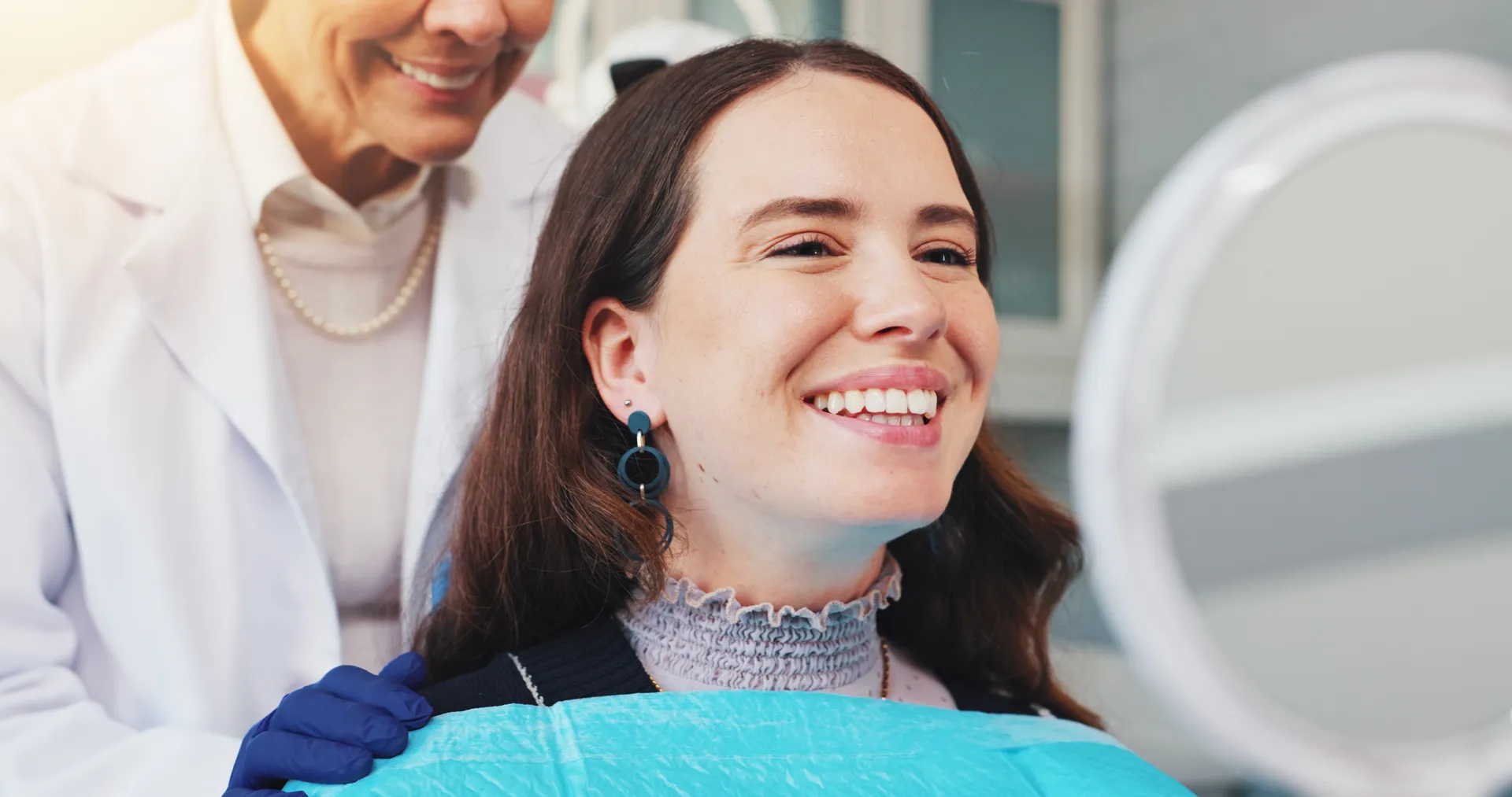Smiling woman looking at her teeth in a mirror while a dental professional wearing gloves stands behind her.