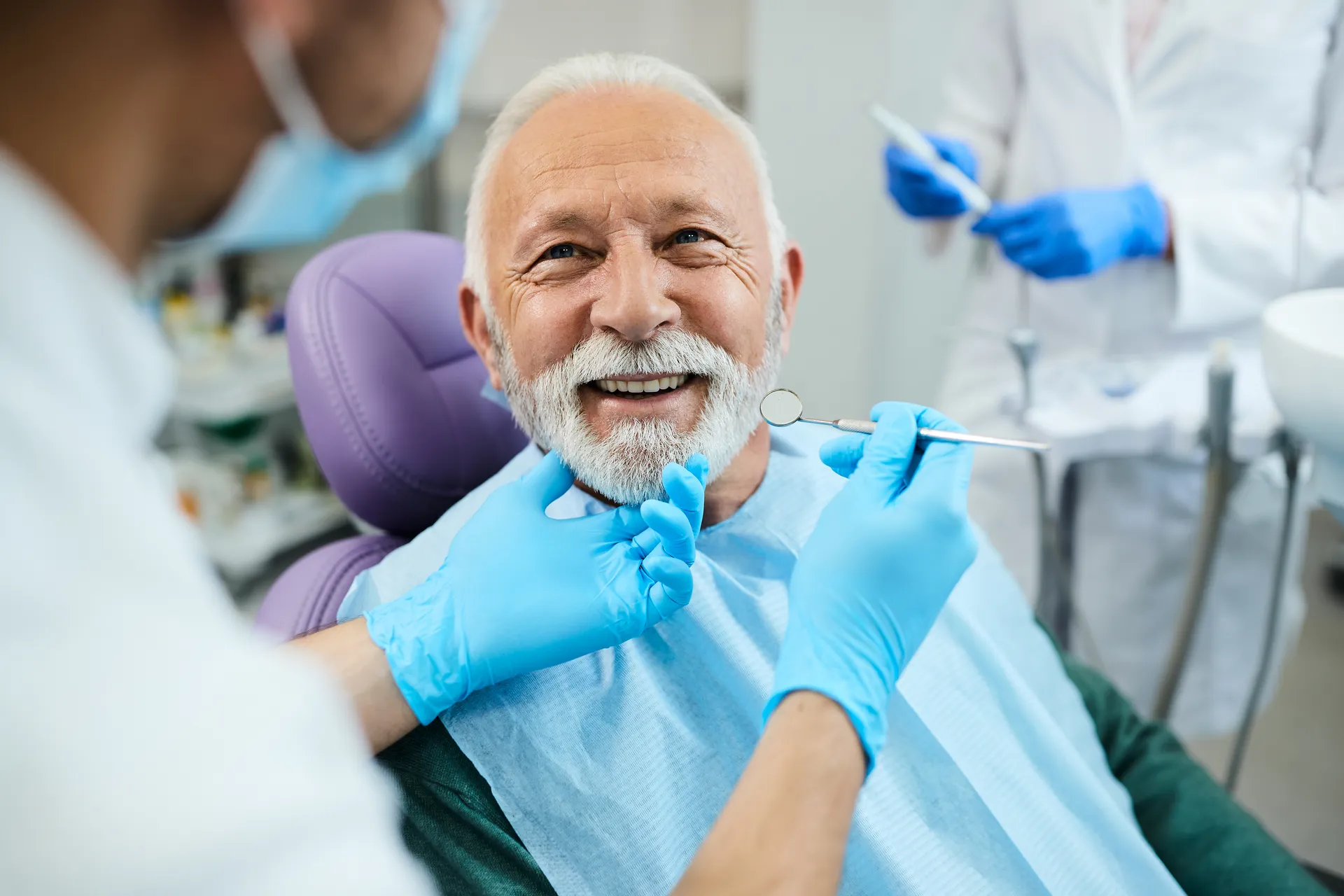 Smiling elderly man with white beard sitting in a dental chair while a dentist wearing blue gloves examines his teeth with a dental mirror.