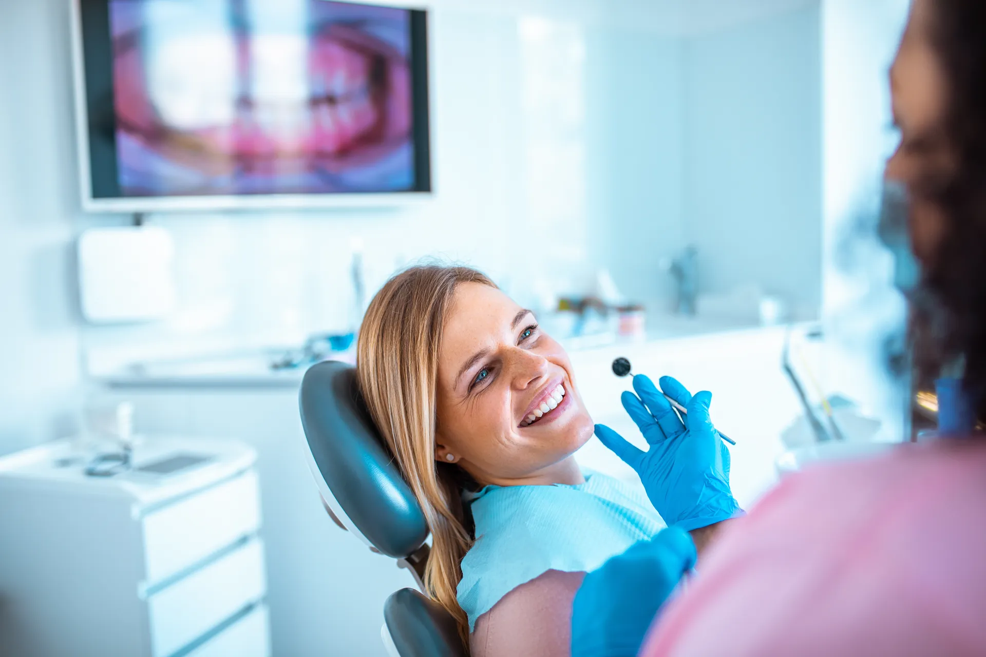 Female dentist wearing blue gloves holding a dental mirror near a smiling woman reclined in a dental chair.