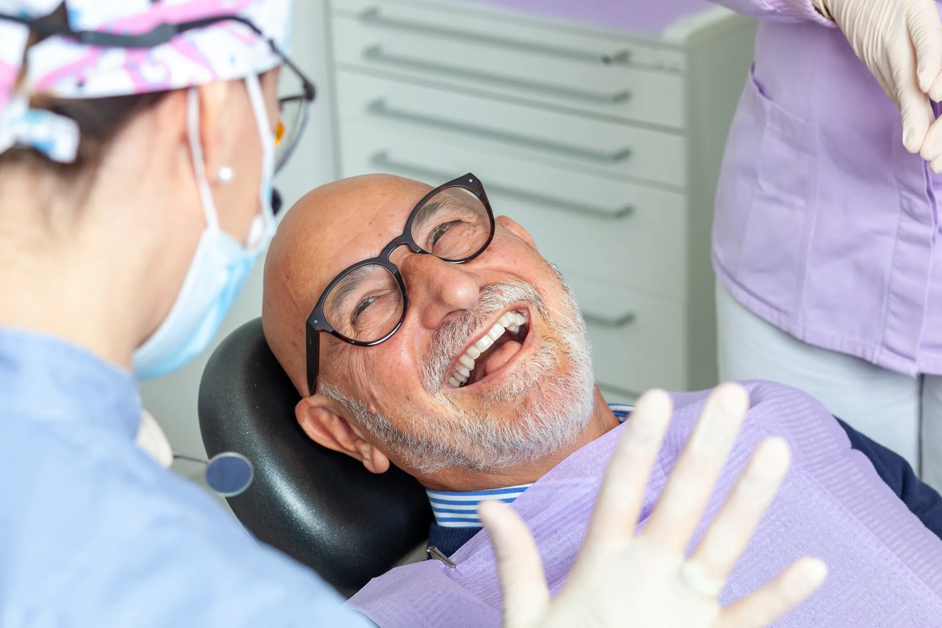 Smiling elderly man with glasses lying in a dental chair during a checkup with a masked dentist.
