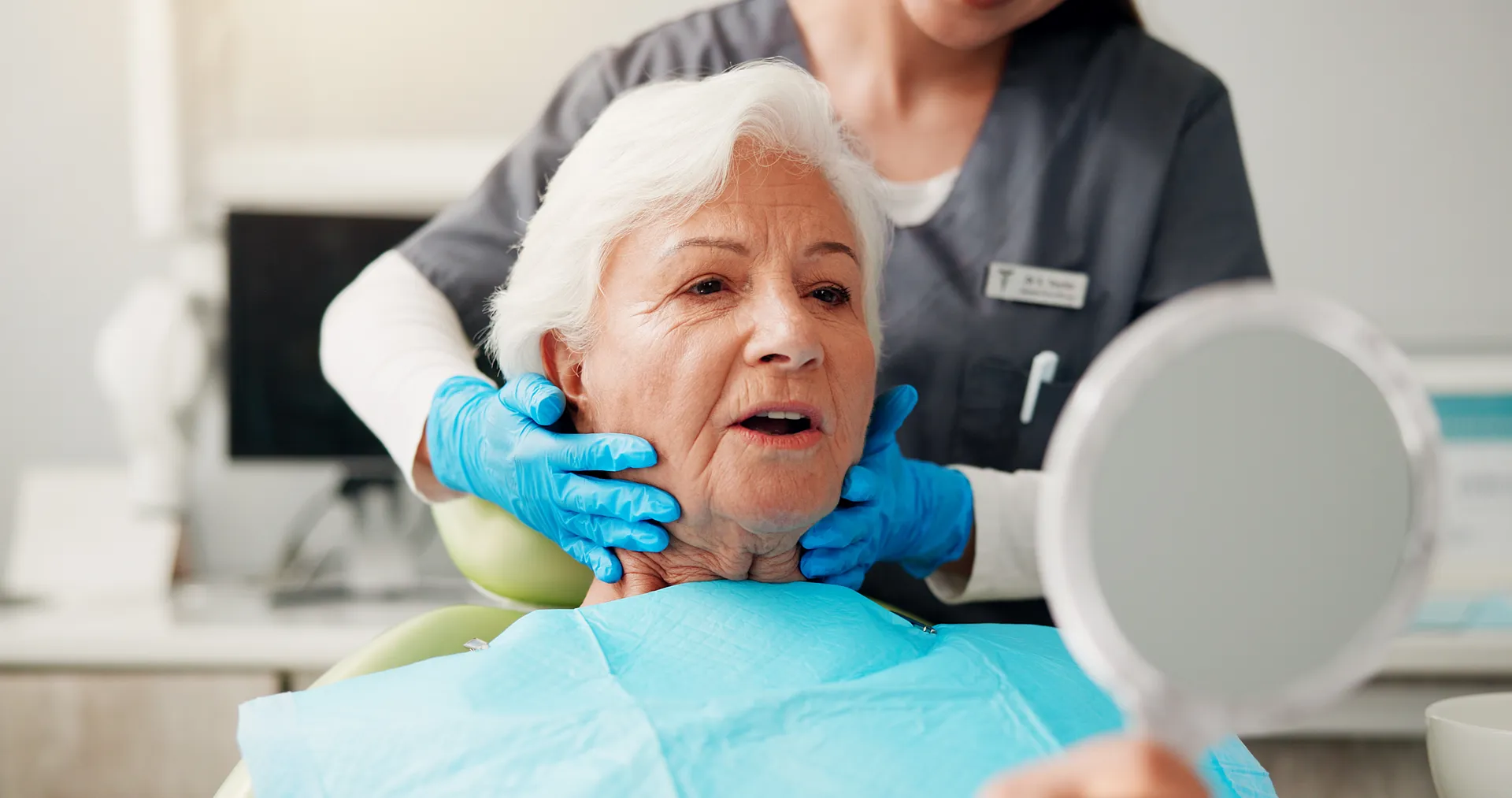 Senior woman sitting in dental chair wearing a bib, with dental professional in gloves examining her jaw while she looks in a hand mirror.