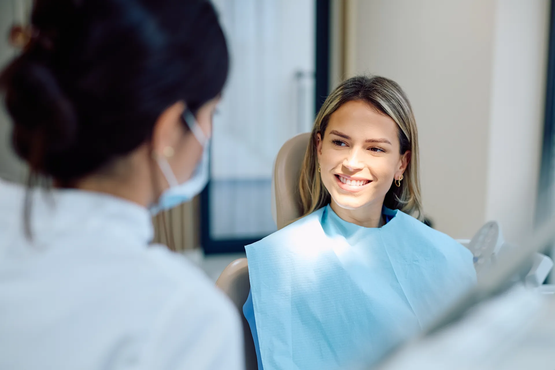 Smiling woman wearing a dental bib sitting in a dental chair, talking with a masked dentist.