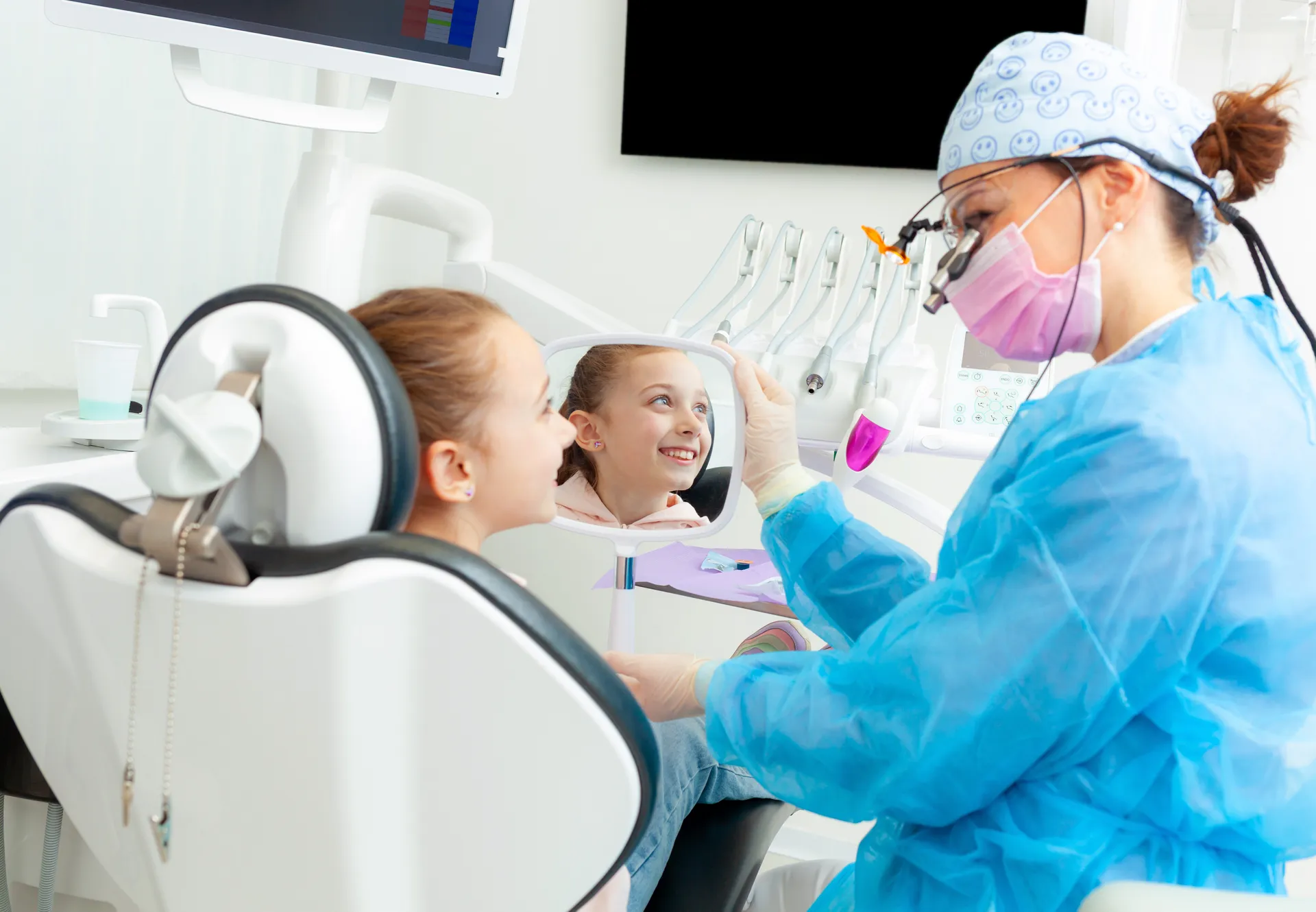 Dentist in blue protective gown showing a young girl her smiling reflection in a handheld mirror at a dental clinic.