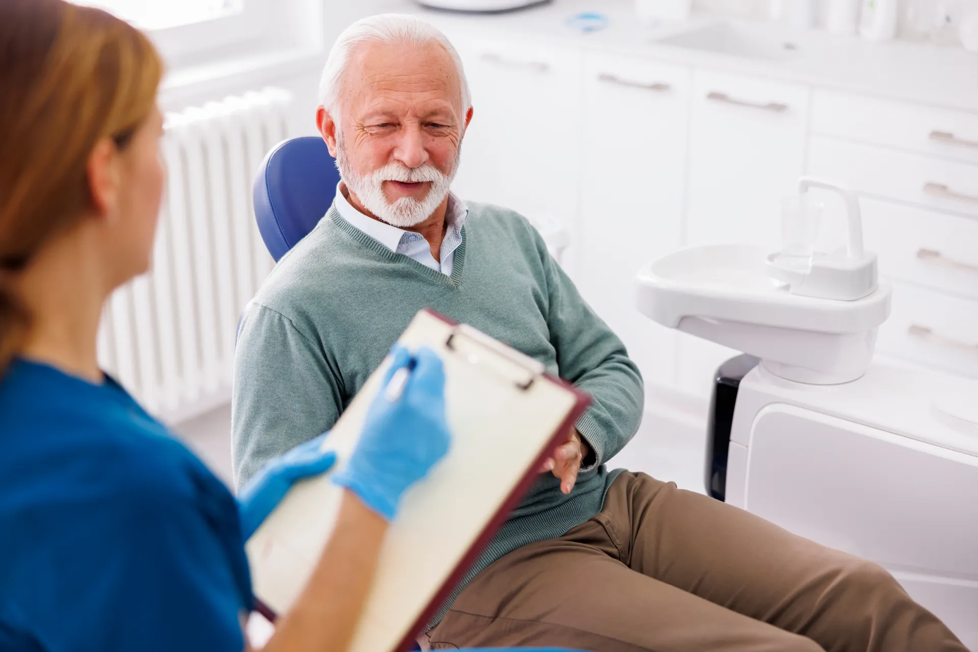 Elderly man sitting in a dental chair talking to a dental professional writing on a clipboard.