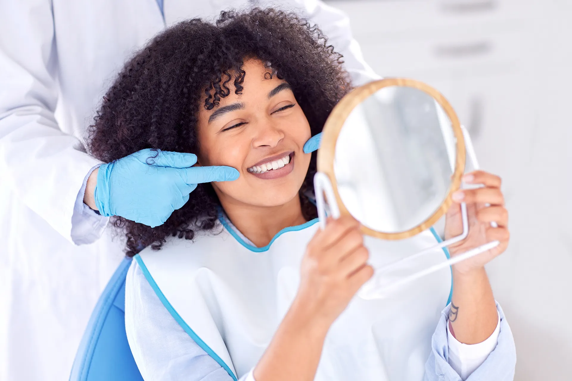 Dentist wearing blue gloves pointing at a smiling woman's teeth while she looks at her reflection in a hand mirror.