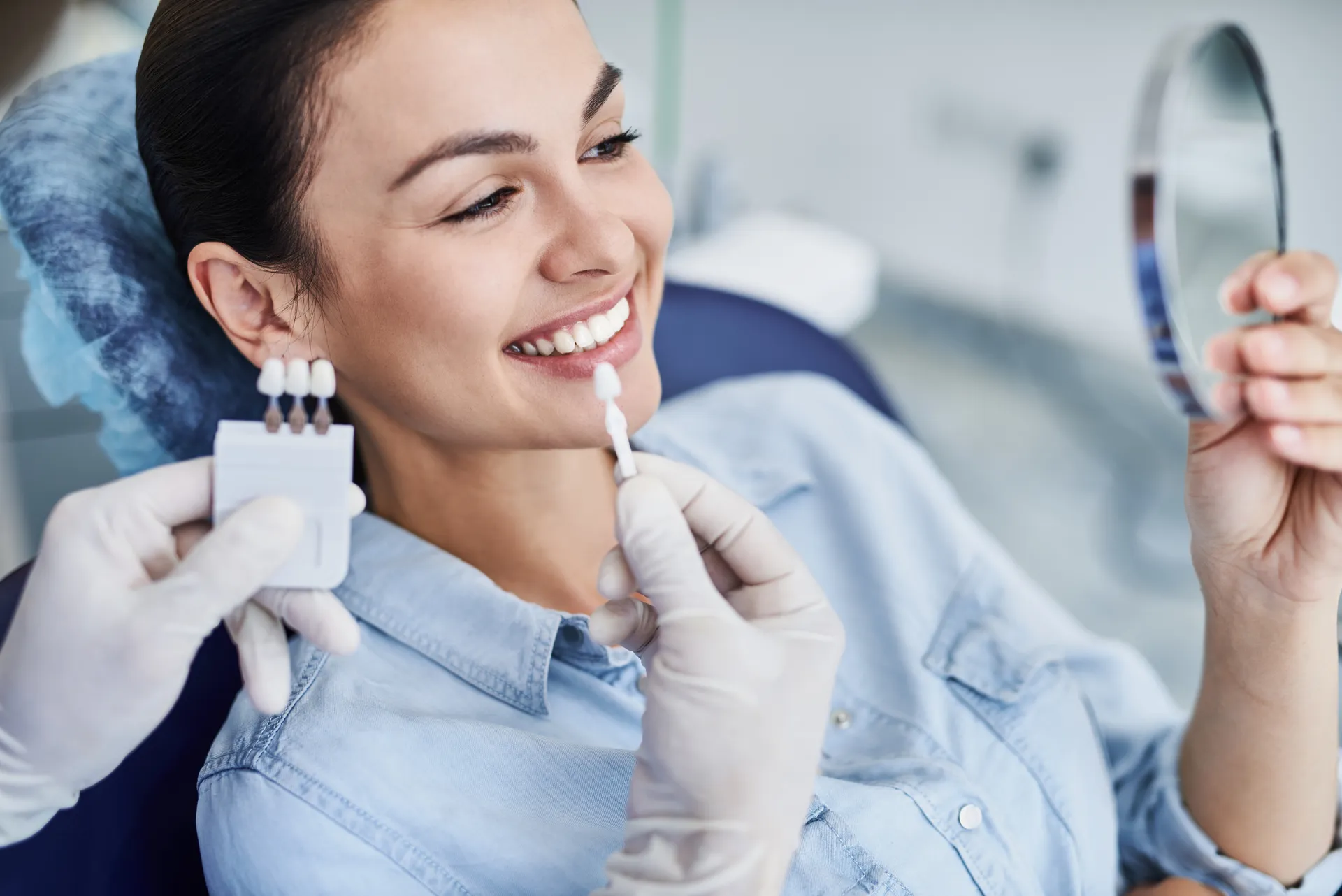 Smiling woman at dentist holding a mirror while dentist compares dental veneer shades to her teeth.