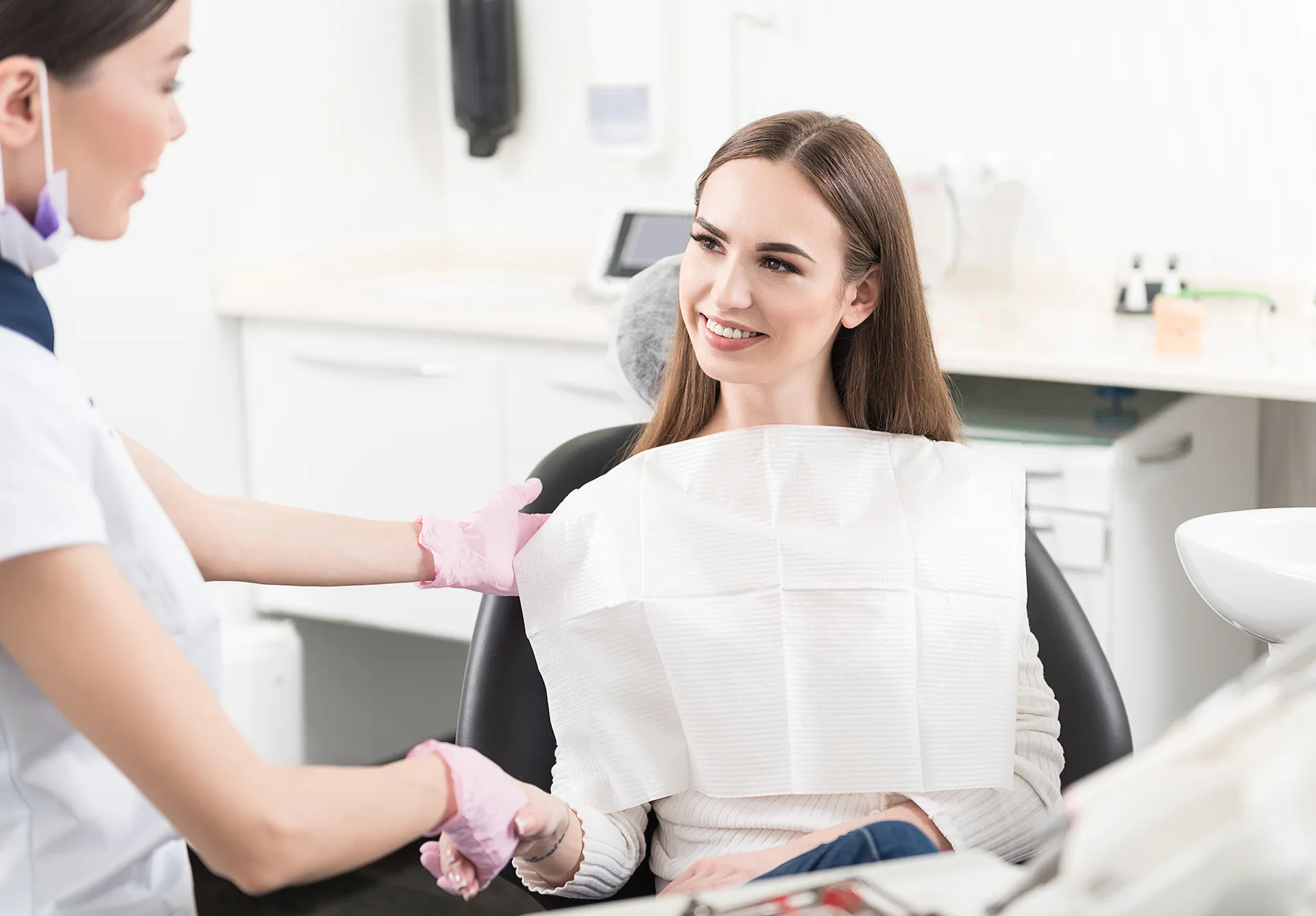 Smiling woman sitting in a dental chair shaking hands with a female dentist wearing pink gloves.