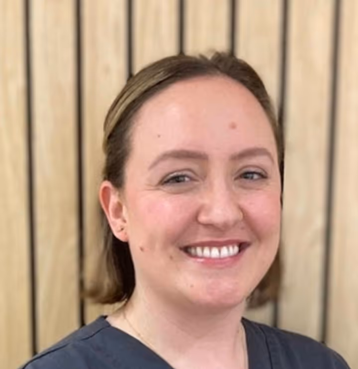 Smiling woman with light skin and brown hair wearing a dark gray shirt standing in front of a wooden slat background.