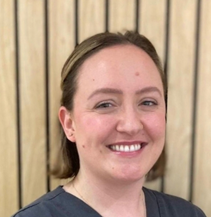 Smiling woman with light skin and brown hair wearing a dark gray shirt standing in front of a wooden slat background.