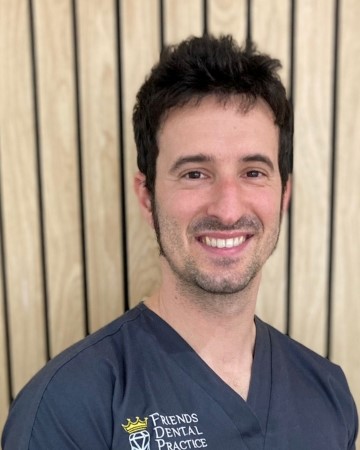 Smiling man wearing a navy dental practice uniform standing in front of a wooden panel background.
