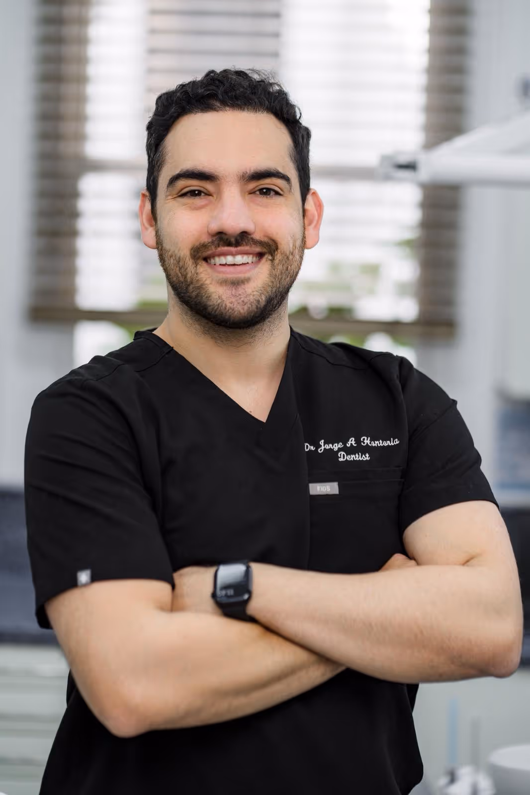 Smiling male dentist in black scrubs with arms crossed standing in a dental office.