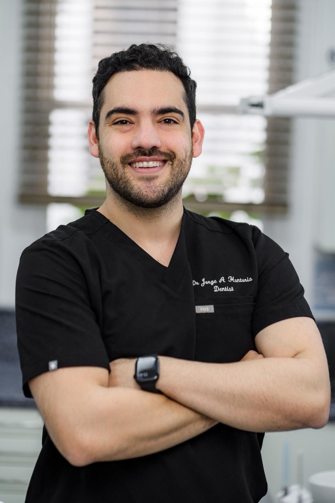 Smiling male dentist in black scrubs with arms crossed standing in a dental office.