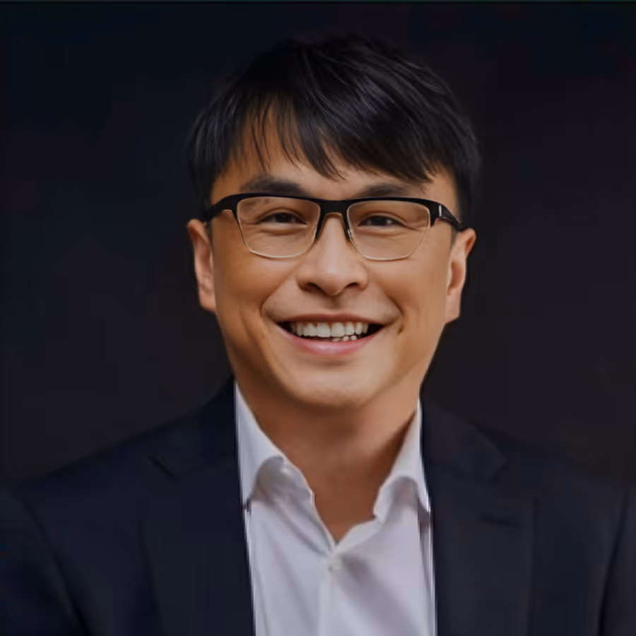 Smiling man with black hair and glasses wearing a white shirt and dark blazer against a dark background.
