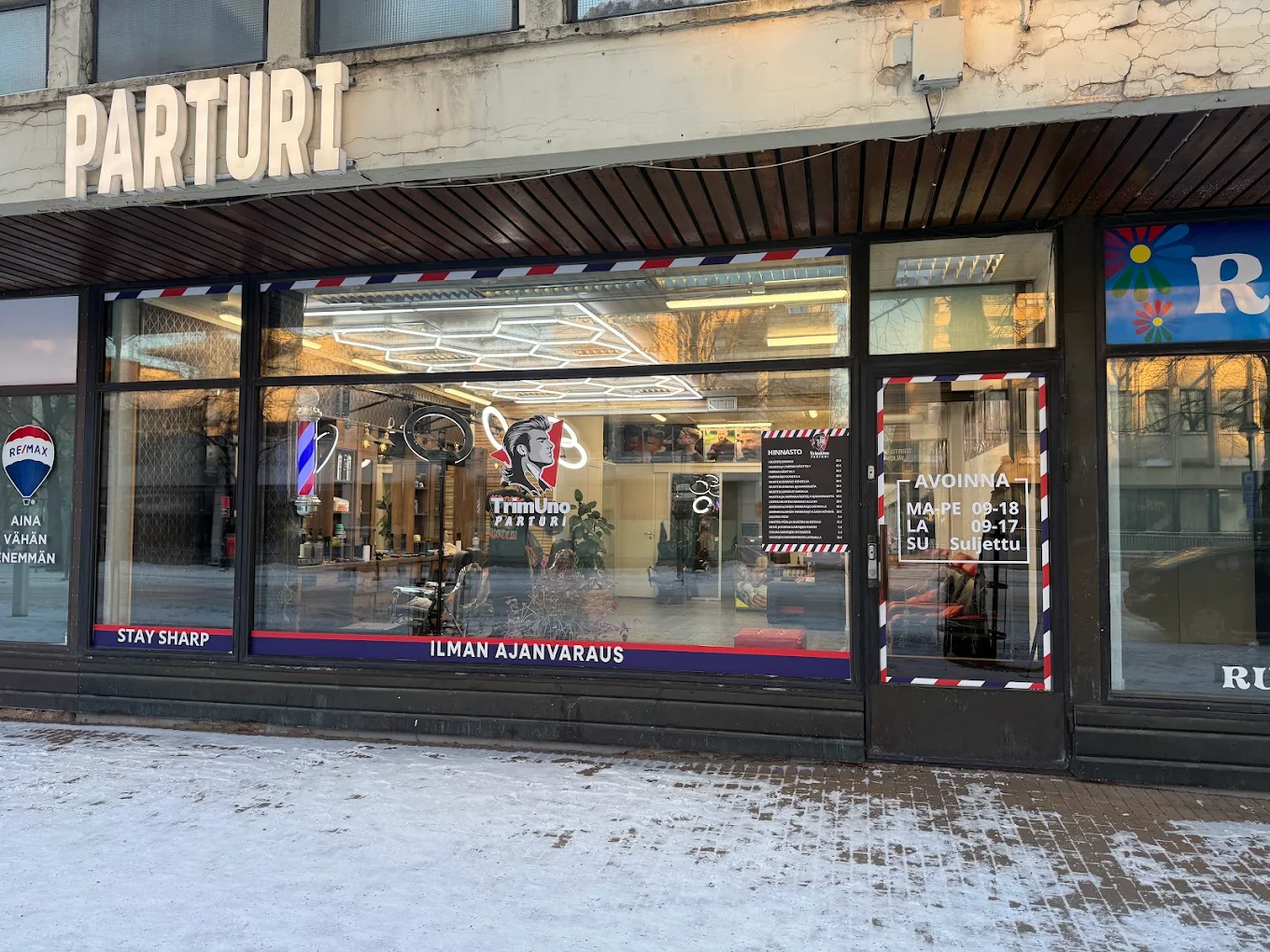 Barbershop front with large glass windows showing interior with barber chairs and neon lights, signage reads 'PARTURI' and operating hours on door.