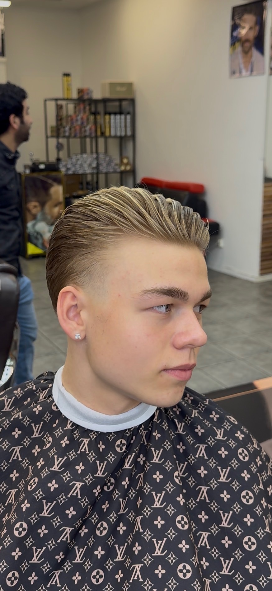 Young man with a slicked-back blonde hairstyle and fade haircut in a barbershop wearing a Louis Vuitton patterned barber cape.