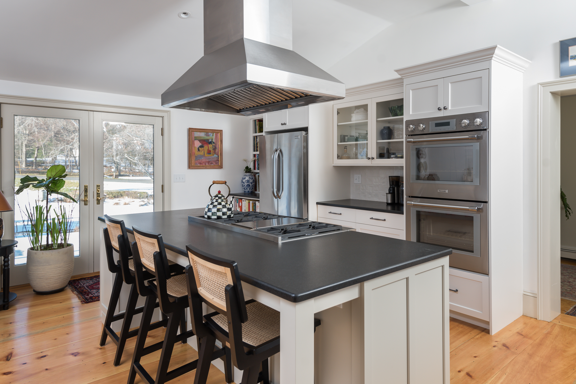 Island with cane counter stools facing French doors, with double wall ovens and glass-front cabinets beyond