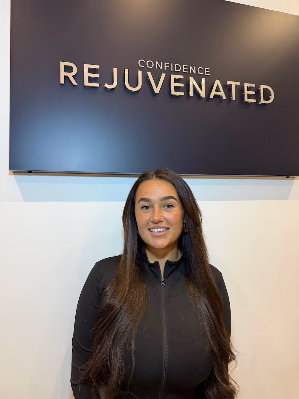 Smiling woman with long brown hair standing in front of a dark sign that reads 'Confidence Rejuvenated'.