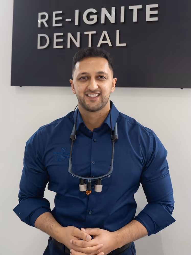 Smiling male dentist in a blue shirt with magnifying loupes around his neck, standing in front of a sign reading 'RE-IGNITE DENTAL'.