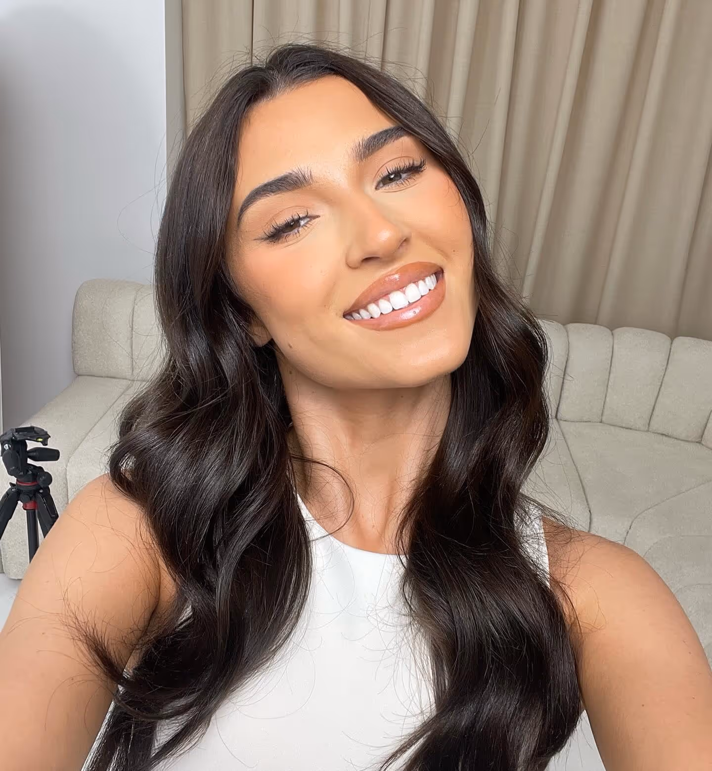 Smiling woman with long dark wavy hair wearing a white top, posing indoors with beige curtains and sofa in background.