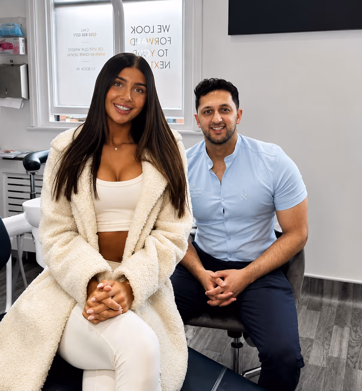 Smiling woman in a white coat and man in light blue shirt seated in a clinic room.