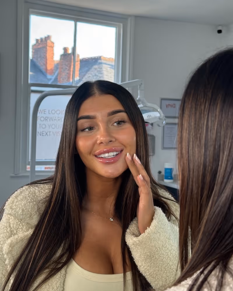 Woman with long dark hair smiling and touching her cheek while looking in a mirror indoors.