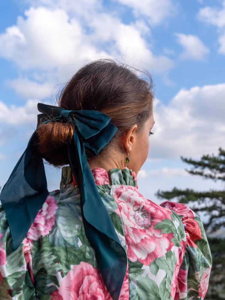 Serena Bifolchi seen from behind, wearing a floral-print dress with a green silk bow, beneath a blue Tuscan sky.