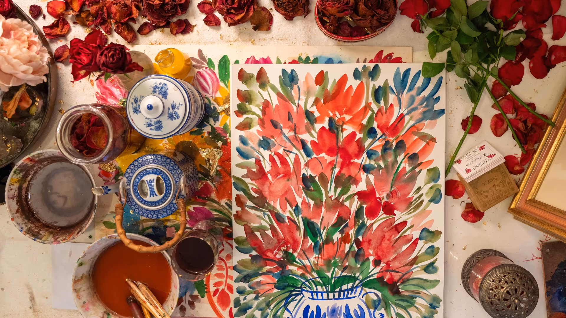 A floral painting surrounded by jars of natural pigments, ceramic bowls, and scattered rose petals in the studio.