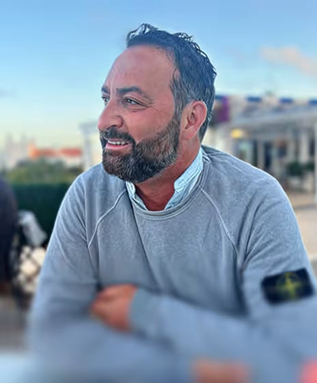 Mann mit Bart und grauem Sweatshirt sitzt entspannt draußen bei klarem Himmel.