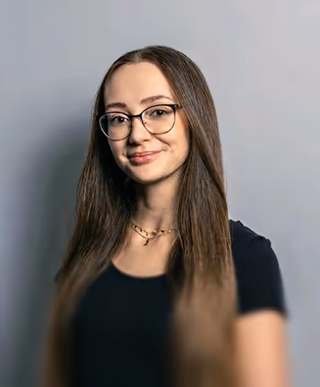 Junge Frau mit langen braunen Haaren, Brille und schwarzem Oberteil vor grauem Hintergrund.