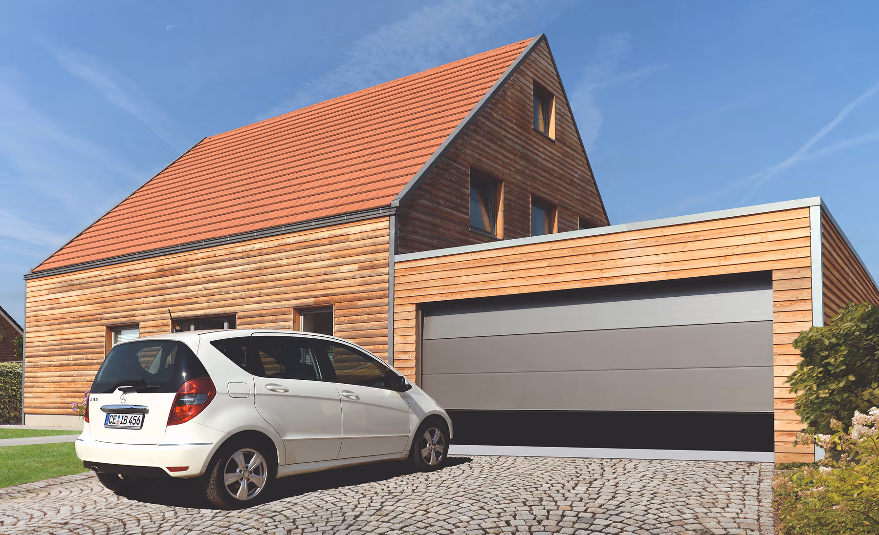 Weißes Auto geparkt auf gepflasterter Einfahrt vor einem modernen Holzhaus mit rotem Dach und Doppelgarage.