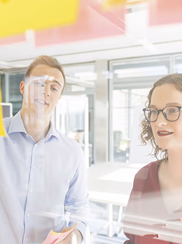 Zwei Personen arbeiten zusammen, schreiben an einem Glasboard mit Haftnotizen in einem modernen Büro.