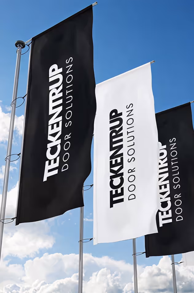 Drei Fahnen mit dem Text ‚Teckentrup Door Solutions‘ wehen vor blauem Himmel mit Wolken.