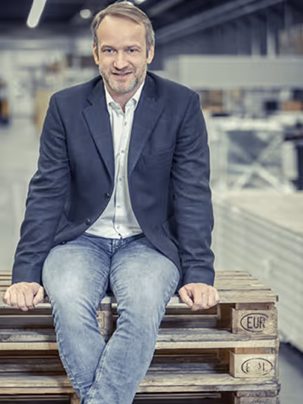 Ein lächelnder Mann mit Sakko und Jeans sitzt auf Holzpaletten in einer Lagerhalle.