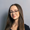 Junge Frau mit langen braunen Haaren und Brille lächelt vor hellgrauem Hintergrund.