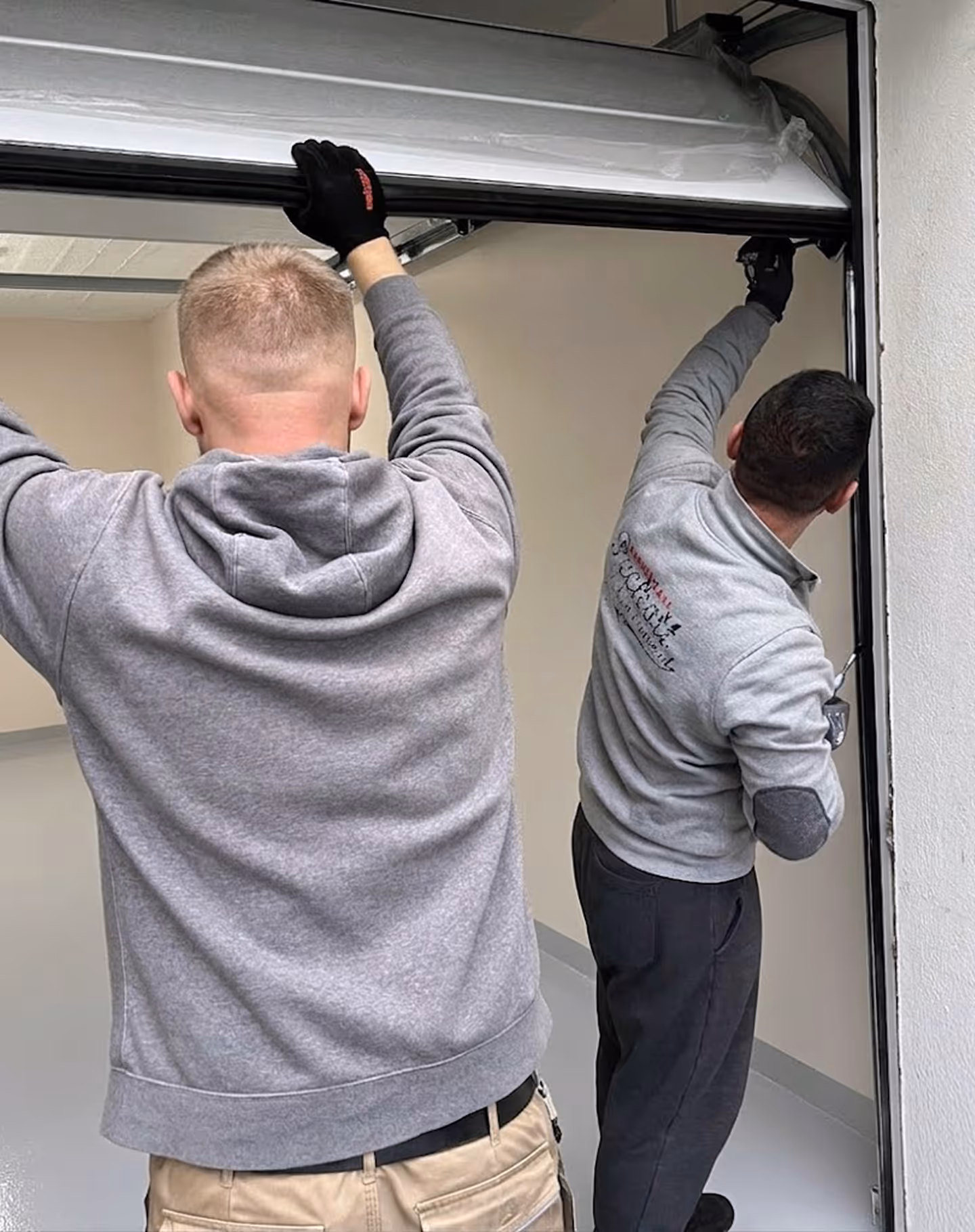 Zwei Männer in grauen Pullovern montieren ein Garagentor in einem leeren Raum.