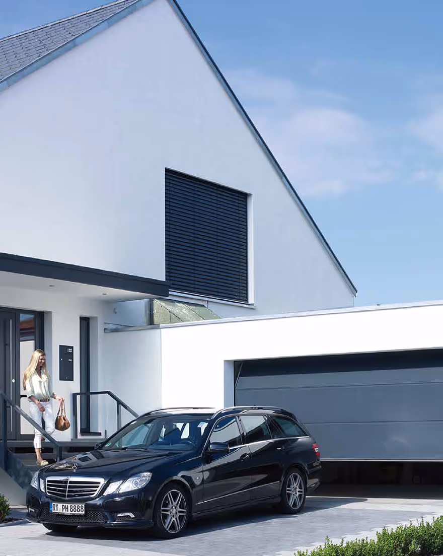 Schwarzer Mercedes-Kombi steht vor einem modernen weißen Haus mit geöffneter Garagentür und einer Frau, die die Eingangstreppe hinuntergeht.