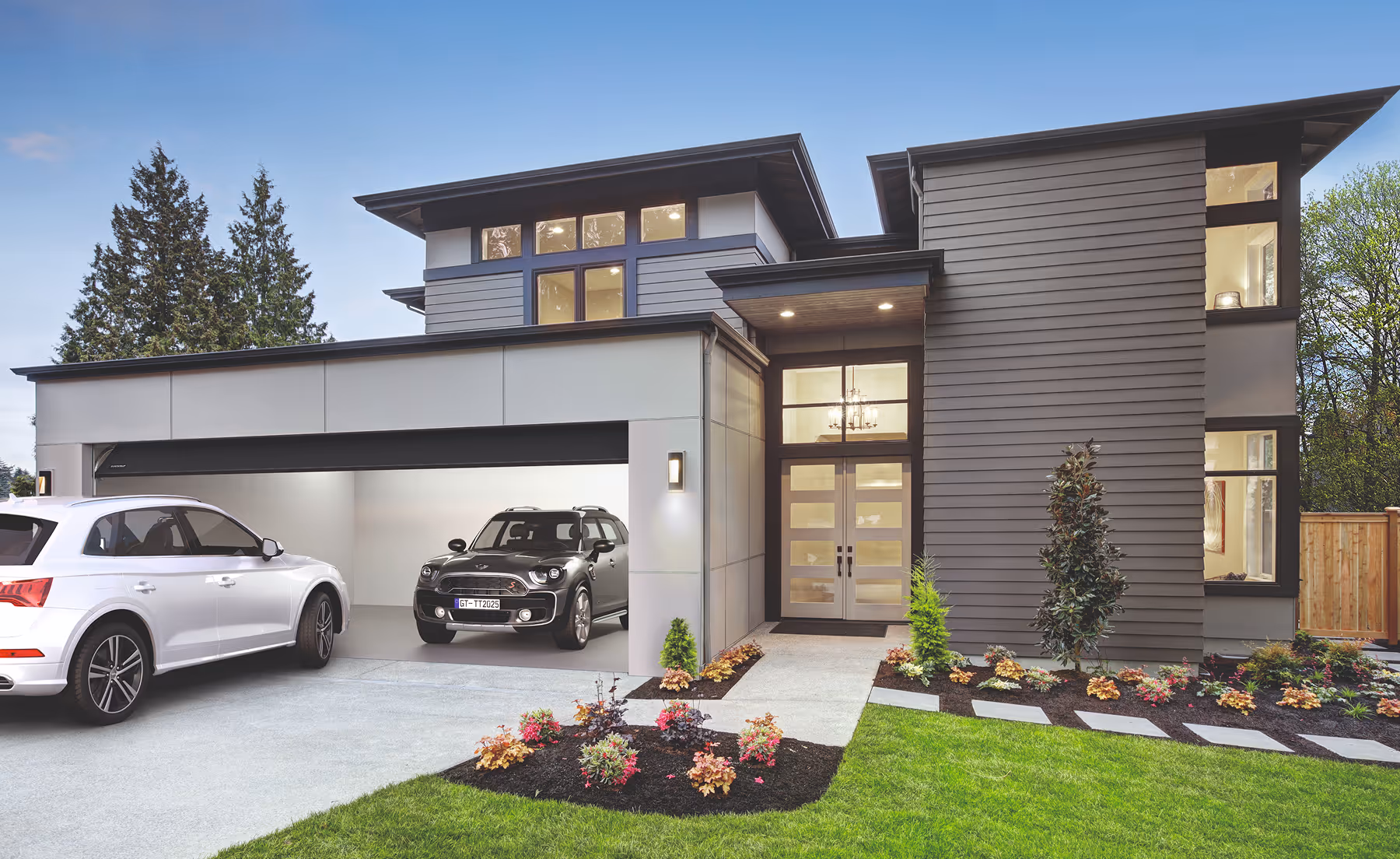 Modernes zweistöckiges Haus mit grauer Fassade, großem Eingangsbereich, Doppelgarage und zwei geparkten Autos, umgeben von grünem Rasen und bunter Bepflanzung.